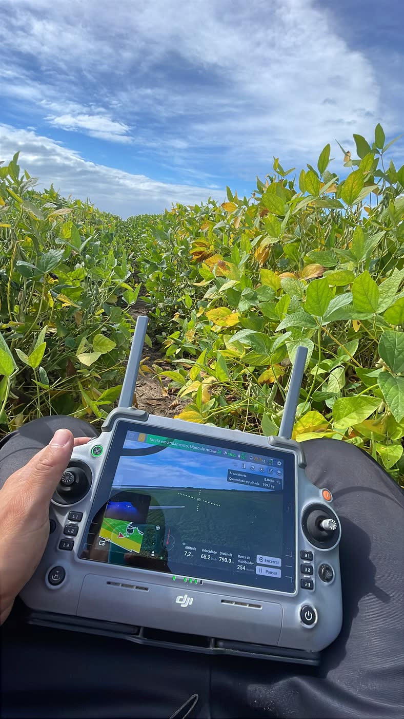 Controle do DJI Agras T100 nas mãos do operador, com a missão em andamento na tela, em lavoura de soja.